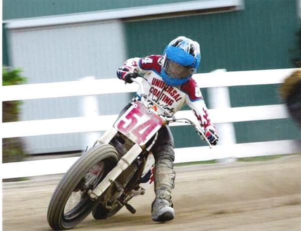 Young Michael leaning his #54 flat-track motorcycle into a dirt turn, wearing a Universal Coating jersey and a blue helmet