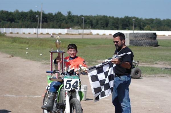 Young Michael on his #54 dirt bike holding a tall trophy while a man waves the checkered flag, before 2005