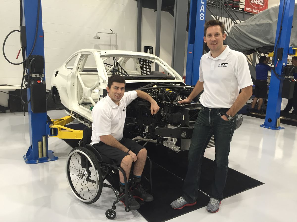 Michael in his wheelchair at a hand-control rig beside a race car on a shop lift, with a team member standing next to him